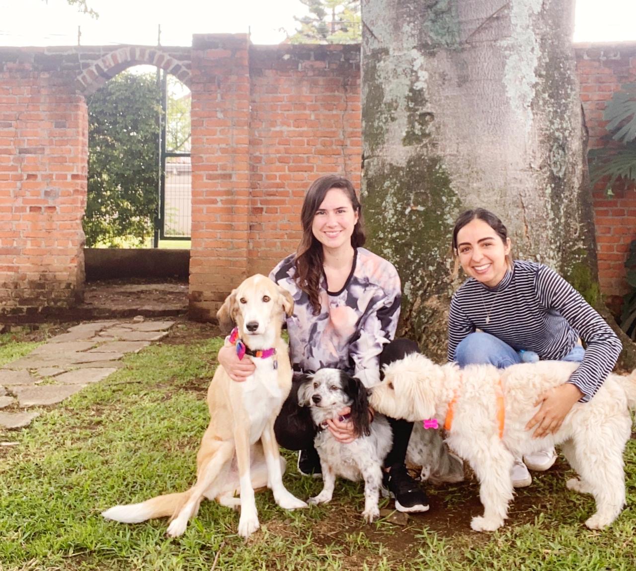 two young brunette women with their 3 rescued dogs. One dog is tall and beige, the other one is medium size with white and black fur, and the third one is small beige and fluffy