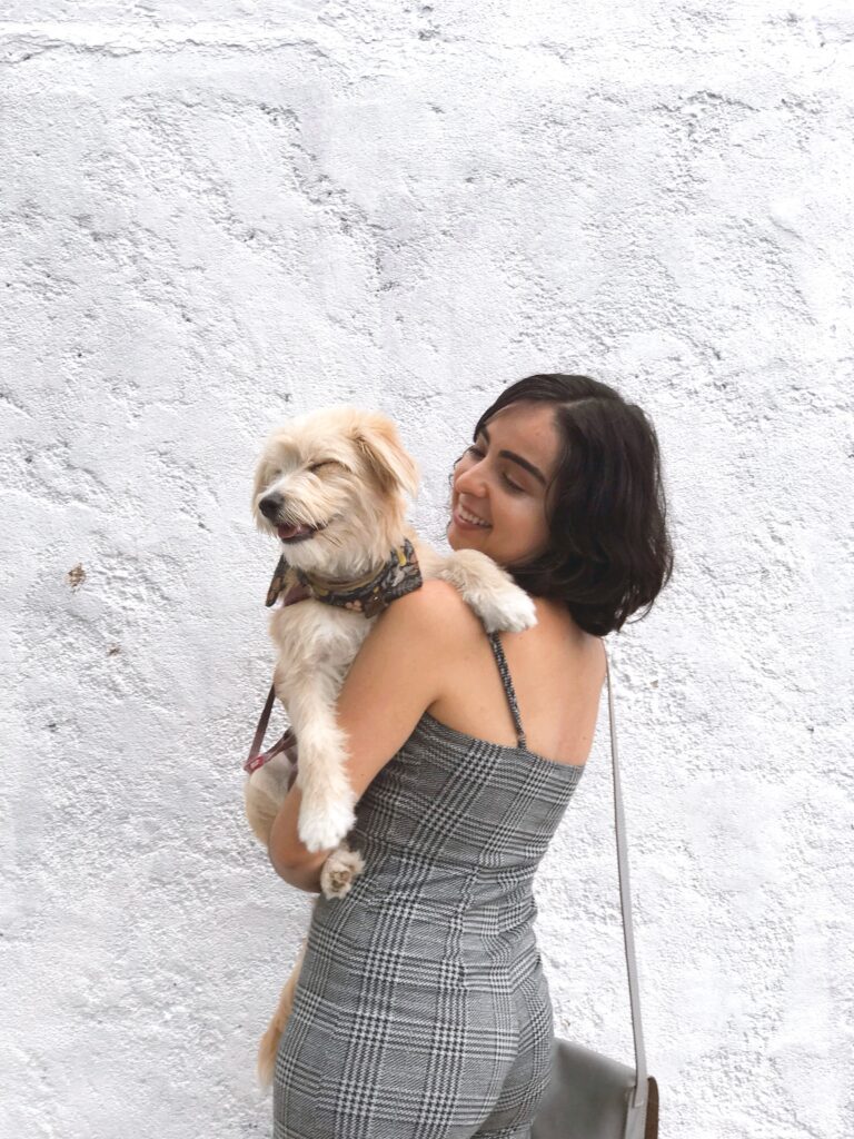 a young woman with short black hair carrying her little beige dog, both are smiling